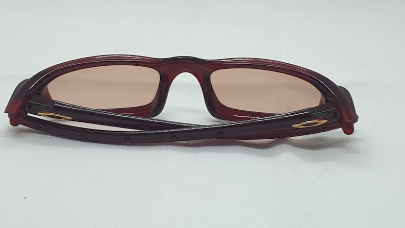 May include: Maroon sunglasses with a rectangular frame and tinted lenses. The arms are folded, revealing a gold-coloured logo on the hinge. The glasses are set against a white background.