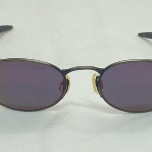 May include: Oval-shaped sunglasses with a dark frame and purple lenses. The temples are thin and wrap around the ears. The nose pads are a light yellow color. The sunglasses are set against a white background.