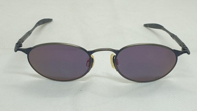 May include: Oval-shaped sunglasses with a dark frame and purple lenses. The temples are thin and wrap around the ears. The nose pads are a light yellow color. The sunglasses are set against a white background.