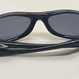 Puede incluir: Gafas de sol negras con lentes grises oscuras. Las gafas tienen una montura y patillas curvas, con un logotipo plateado en las patillas. Las lentes son de forma ovalada y protegen del sol.