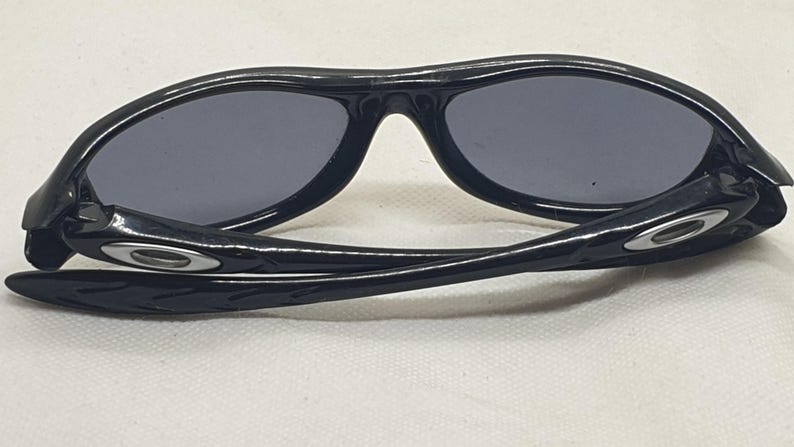 Puede incluir: Gafas de sol negras con lentes grises oscuras. Las gafas tienen una montura y patillas curvas, con un logotipo plateado en las patillas. Las lentes son de forma ovalada y protegen del sol.