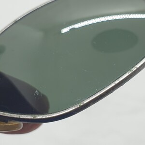Puede incluir: Primer plano de gafas de sol vintage con lentes verdes y montura plateada. Las gafas tienen una forma de aviador cl&aacute;sica, con un peque&ntilde;o acento dorado en la montura. Las lentes est&aacute;n ligeramente rayadas.