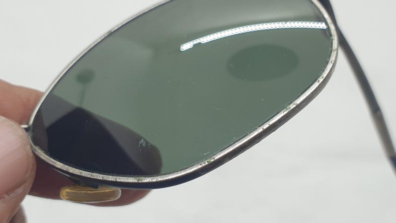 Puede incluir: Primer plano de gafas de sol vintage con lentes verdes y montura plateada. Las gafas tienen una forma de aviador cl&aacute;sica, con un peque&ntilde;o acento dorado en la montura. Las lentes est&aacute;n ligeramente rayadas.