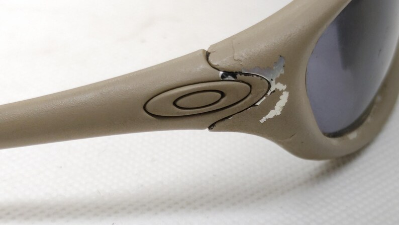 Puede incluir: Primer plano de gafas de sol color beige. La montura muestra desgaste con pintura desconchada. Las lentes son de color gris oscuro. El logotipo de Oakley es visible en la patilla.