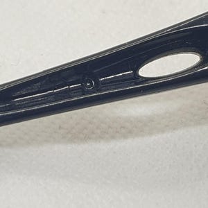 Puede incluir: Primer plano de una patilla de gafas negra y brillante. La patilla presenta un dise&ntilde;o elegante y aerodin&aacute;mico con una peque&ntilde;a abertura ovalada y una sutil ranura. La superficie refleja la luz, resaltando su textura suave.