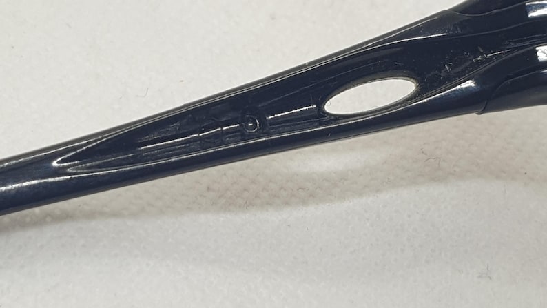 Puede incluir: Primer plano de una patilla de gafas negra y brillante. La patilla presenta un dise&ntilde;o elegante y aerodin&aacute;mico con una peque&ntilde;a abertura ovalada y una sutil ranura. La superficie refleja la luz, resaltando su textura suave.