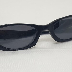 May include: Black sunglasses with a sleek, curved design. The sunglasses have dark lenses and a black frame. The left arm of the sunglasses has a small, silver-coloured metal detail. The sunglasses are set against a white background.