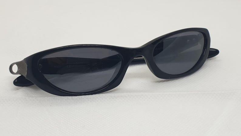 May include: Black sunglasses with a sleek, curved design. The sunglasses have dark lenses and a black frame. The left arm of the sunglasses has a small, silver-coloured metal detail. The sunglasses are set against a white background.