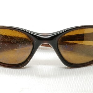 Puede incluir: Gafas de sol marrones con montura curvada y lentes &aacute;mbar. Las gafas tienen un dise&ntilde;o retro cl&aacute;sico, con una montura marr&oacute;n oscuro y una forma sutilmente curvada. Las lentes son de color &aacute;mbar c&aacute;lido.