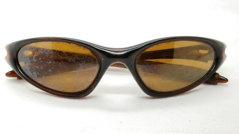 Puede incluir: Gafas de sol marrones con montura curvada y lentes &aacute;mbar. Las gafas tienen un dise&ntilde;o retro cl&aacute;sico, con una montura marr&oacute;n oscuro y una forma sutilmente curvada. Las lentes son de color &aacute;mbar c&aacute;lido.