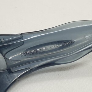 May include: Close-up of the arm of a pair of gray sunglasses. The text "MADE IN USA" is embossed on the arm. The sunglasses appear to be made of a translucent plastic material.