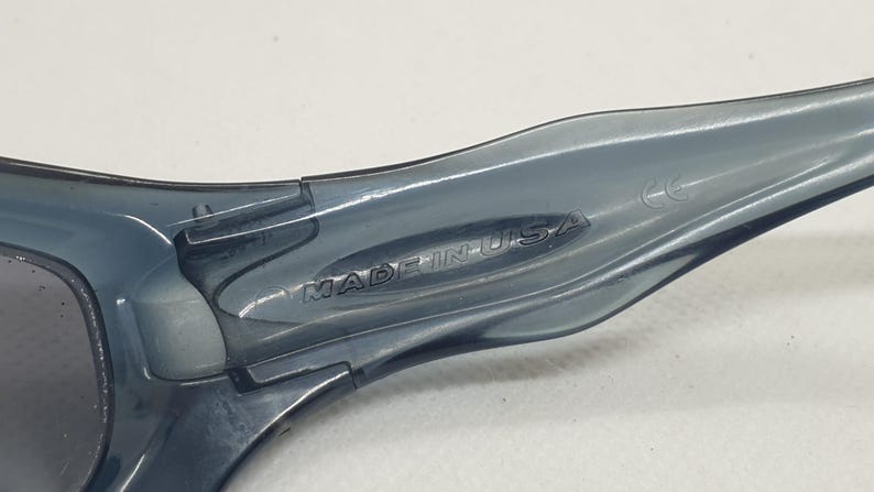 May include: Close-up of the arm of a pair of gray sunglasses. The text "MADE IN USA" is embossed on the arm. The sunglasses appear to be made of a translucent plastic material.