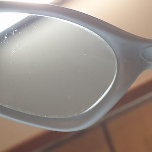 K&ouml;nnte beinhalten: Nahaufnahme einer grauen Sonnenbrille mit ovalen Gl&auml;sern. Der Rahmen hat eine matte Oberfl&auml;che und ein dezentes Logo-Detail in der N&auml;he des Scharniers. Die Gl&auml;ser scheinen reflektierend zu sein, und das Gesamtdesign ist elegant und modern.