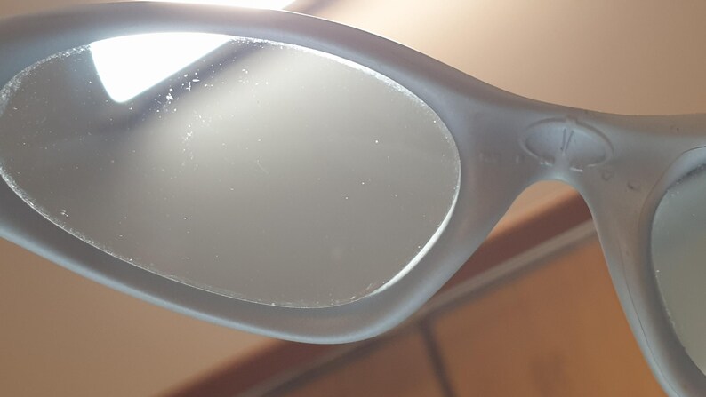 K&ouml;nnte beinhalten: Nahaufnahme einer grauen Sonnenbrille mit ovalen Gl&auml;sern. Der Rahmen hat eine matte Oberfl&auml;che und ein dezentes Logo-Detail in der N&auml;he des Scharniers. Die Gl&auml;ser scheinen reflektierend zu sein, und das Gesamtdesign ist elegant und modern.