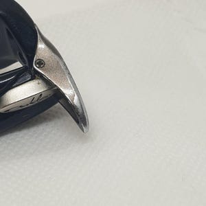 May include: Close-up of black sunglasses with a silver hinge. The sunglasses have dark lenses and a sleek design. The hinge features a small screw and a brand name.