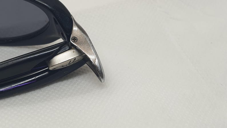 May include: Close-up of black sunglasses with a silver hinge. The sunglasses have dark lenses and a sleek design. The hinge features a small screw and a brand name.