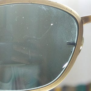 May include: Close-up of Ray-Ban sunglasses with a gold-colored frame and dark lenses. The Ray-Ban logo is visible on the lens. The sunglasses are held by a person's fingers.