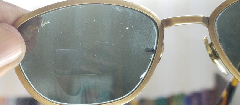 May include: Close-up of Ray-Ban sunglasses with a gold-colored frame and dark lenses. The Ray-Ban logo is visible on the lens. The sunglasses are held by a person's fingers.