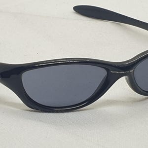 Puede incluir: Gafas de sol negras con lentes de color gris oscuro. Las gafas de sol tienen un dise&ntilde;o elegante y curvado con un acabado brillante. La montura presenta el logotipo de Oakley en las patillas. Estas gafas est&aacute;n dise&ntilde;adas para la protecci&oacute;n ocular.