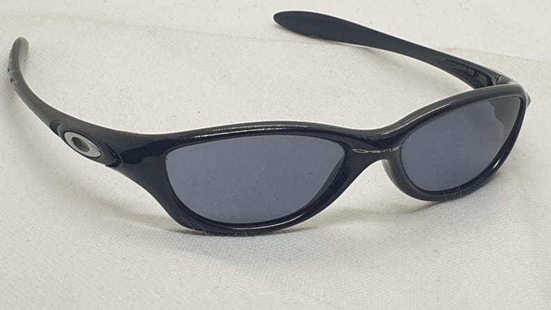 Puede incluir: Gafas de sol negras con lentes de color gris oscuro. Las gafas de sol tienen un dise&ntilde;o elegante y curvado con un acabado brillante. La montura presenta el logotipo de Oakley en las patillas. Estas gafas est&aacute;n dise&ntilde;adas para la protecci&oacute;n ocular.