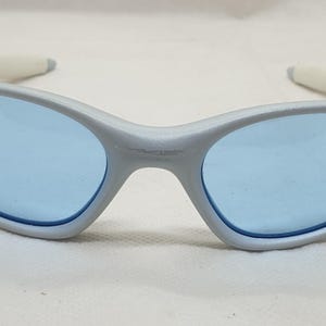 K&ouml;nnte beinhalten: Silberfarbene Sonnenbrille mit hellblauen Gl&auml;sern. Die Sonnenbrille hat ein geschwungenes Design mit wei&szlig;en B&uuml;gelenden. Die Gl&auml;ser sind oval und mit einem dunkelblauen Rand versehen. Die Sonnenbrille liegt auf einer wei&szlig;en Oberfl&auml;che.