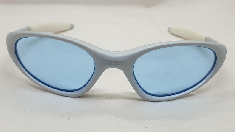 K&ouml;nnte beinhalten: Silberfarbene Sonnenbrille mit hellblauen Gl&auml;sern. Die Sonnenbrille hat ein geschwungenes Design mit wei&szlig;en B&uuml;gelenden. Die Gl&auml;ser sind oval und mit einem dunkelblauen Rand versehen. Die Sonnenbrille liegt auf einer wei&szlig;en Oberfl&auml;che.