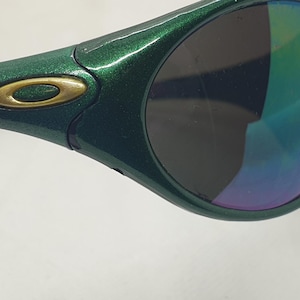 May include: Close-up of a pair of green sunglasses with a metallic finish. The lenses have a gradient of colors, including blue, green, and purple. The frame features a gold-colored logo.