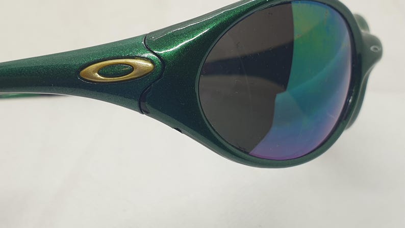 May include: Close-up of a pair of green sunglasses with a metallic finish. The lenses have a gradient of colors, including blue, green, and purple. The frame features a gold-colored logo.