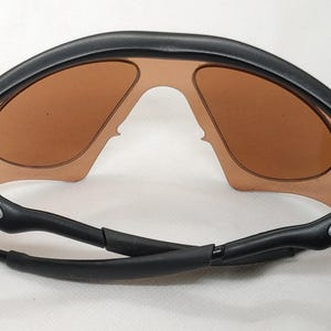 May include: Black sunglasses with brown tinted lenses. The sunglasses have a curved, wraparound design with a black frame and arms. The Oakley logo is visible on the arms. The lenses are a warm, light brown colour.