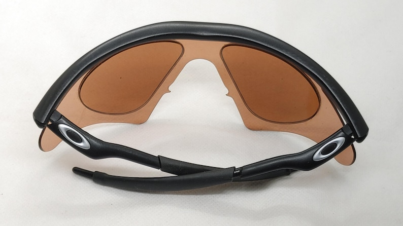 May include: Black sunglasses with brown tinted lenses. The sunglasses have a curved, wraparound design with a black frame and arms. The Oakley logo is visible on the arms. The lenses are a warm, light brown colour.