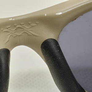 Puede incluir: Primer plano de gafas de sol con montura beige y lentes grises. La montura tiene un peque&ntilde;o emblema decorativo cerca del puente. Las almohadillas nasales son negras.