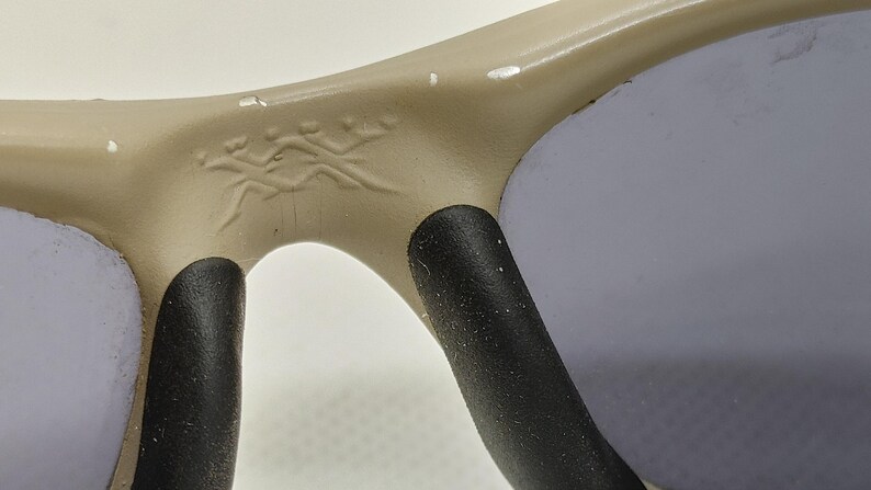 Puede incluir: Primer plano de gafas de sol con montura beige y lentes grises. La montura tiene un peque&ntilde;o emblema decorativo cerca del puente. Las almohadillas nasales son negras.