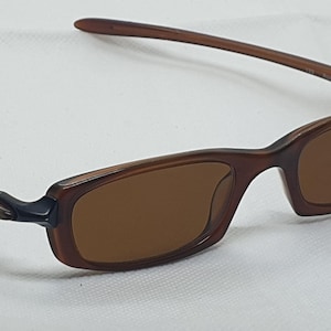 Pode incluir: &Oacute;culos de sol retangulares castanhos com lentes castanhas. As arma&ccedil;&otilde;es s&atilde;o de um tom castanho mais escuro, com um design elegante e moderno. As hastes s&atilde;o finas e curvam-se suavemente em dire&ccedil;&atilde;o &agrave;s orelhas. Os &oacute;culos est&atilde;o sobre um fundo branco.