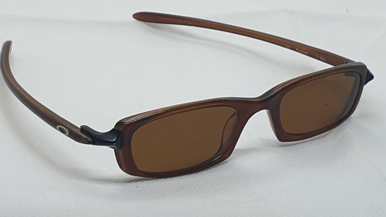 Pode incluir: &Oacute;culos de sol retangulares castanhos com lentes castanhas. As arma&ccedil;&otilde;es s&atilde;o de um tom castanho mais escuro, com um design elegante e moderno. As hastes s&atilde;o finas e curvam-se suavemente em dire&ccedil;&atilde;o &agrave;s orelhas. Os &oacute;culos est&atilde;o sobre um fundo branco.