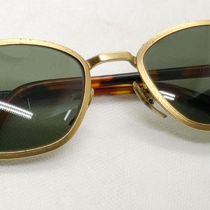 May include: Gold-framed sunglasses with green lenses and a tortoise shell pattern on the arms. The Ray-Ban logo is visible on the left lens. The sunglasses have a classic, vintage design.