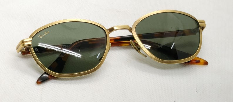May include: Gold-framed sunglasses with green lenses and a tortoise shell pattern on the arms. The Ray-Ban logo is visible on the left lens. The sunglasses have a classic, vintage design.