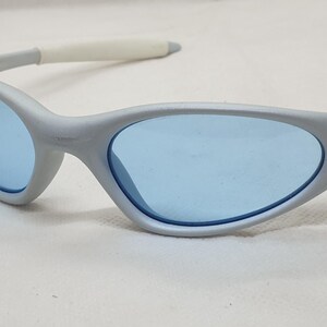 K&ouml;nnte beinhalten: Silbergraue Sonnenbrille mit hellblauen Gl&auml;sern. Die Sonnenbrille hat ein geschwungenes, stromlinienf&ouml;rmiges Design mit wei&szlig;en B&uuml;gelenden. Das Oakley-Logo ist auf der Seite des Rahmens sichtbar. Geeignet f&uuml;r Outdoor-Aktivit&auml;ten.