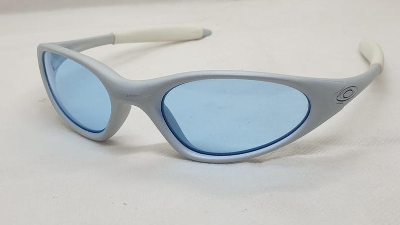 K&ouml;nnte beinhalten: Silbergraue Sonnenbrille mit hellblauen Gl&auml;sern. Die Sonnenbrille hat ein geschwungenes, stromlinienf&ouml;rmiges Design mit wei&szlig;en B&uuml;gelenden. Das Oakley-Logo ist auf der Seite des Rahmens sichtbar. Geeignet f&uuml;r Outdoor-Aktivit&auml;ten.