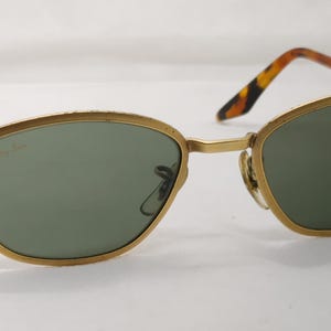 May include: Gold-rimmed sunglasses with green lenses. The frames have a classic oval shape. The temples are a tortoiseshell pattern. The sunglasses are set against a white background.
