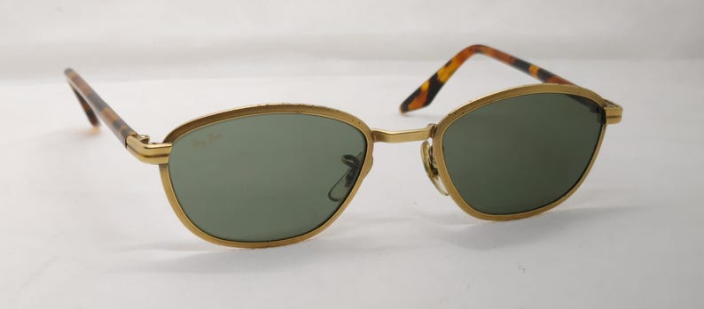 May include: Gold-rimmed sunglasses with green lenses. The frames have a classic oval shape. The temples are a tortoiseshell pattern. The sunglasses are set against a white background.