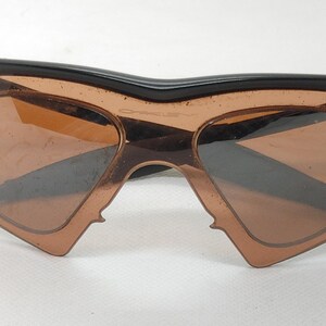 May include: Unusual Oakley sunglasses featuring a black frame and brown lenses. The lenses are heart-shaped. The sunglasses have a distinctive, angular shape, and are set against a white background.
