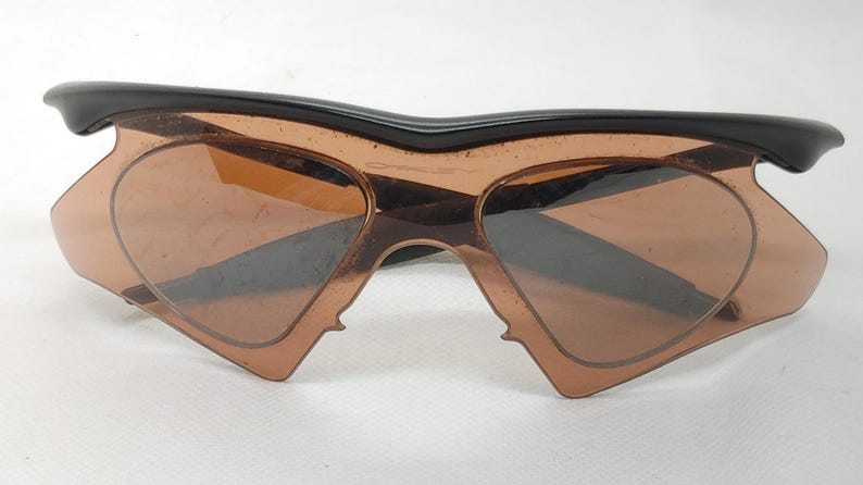 May include: Unusual Oakley sunglasses featuring a black frame and brown lenses. The lenses are heart-shaped. The sunglasses have a distinctive, angular shape, and are set against a white background.