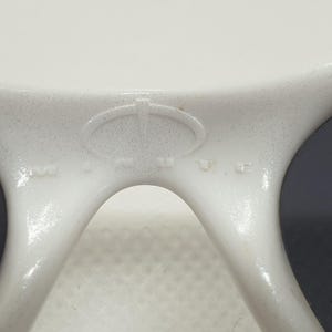 Puede incluir: Primer plano de gafas de sol blancas con lentes oscuras. La montura tiene un acabado texturizado y un logotipo en el puente. Las gafas de sol est&aacute;n dise&ntilde;adas para proteger los ojos de la luz solar.