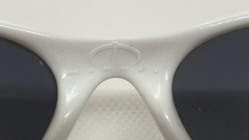 Puede incluir: Primer plano de gafas de sol blancas con lentes oscuras. La montura tiene un acabado texturizado y un logotipo en el puente. Las gafas de sol est&aacute;n dise&ntilde;adas para proteger los ojos de la luz solar.