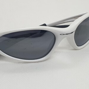 Puede incluir: Gafas de sol blancas con lentes grises oscuras. Las gafas de sol tienen un dise&ntilde;o elegante y curvado con un peque&ntilde;o logotipo en el lateral. La montura es de color blanco brillante y las lentes son de color gris oscuro.