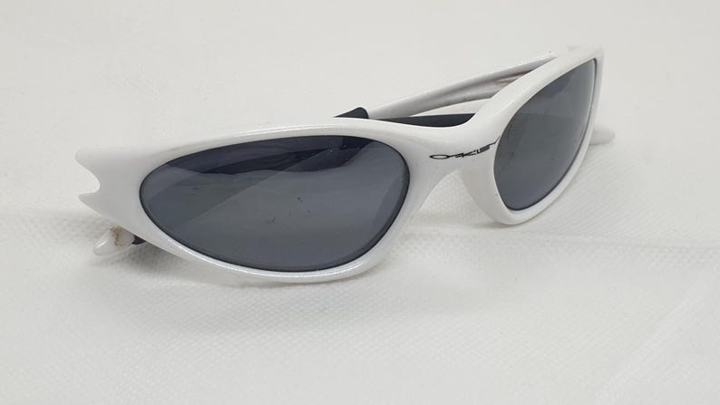 Puede incluir: Gafas de sol blancas con lentes grises oscuras. Las gafas de sol tienen un dise&ntilde;o elegante y curvado con un peque&ntilde;o logotipo en el lateral. La montura es de color blanco brillante y las lentes son de color gris oscuro.