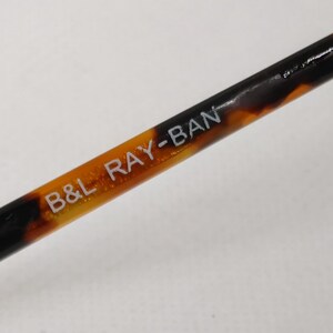 May include: Close-up of a Ray-Ban eyeglass arm with a tortoiseshell pattern. The text "B&L RAY-BAN" is printed in white on the arm. The arm is a mix of brown, black, and amber colors.