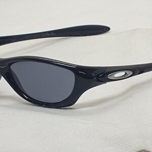 Puede incluir: Gafas de sol negras con lentes grises. Las gafas tienen una montura y patillas curvas, con un peque&ntilde;o emblema plateado en las patillas. Las lentes son de color gris oscuro y la montura es negra brillante.