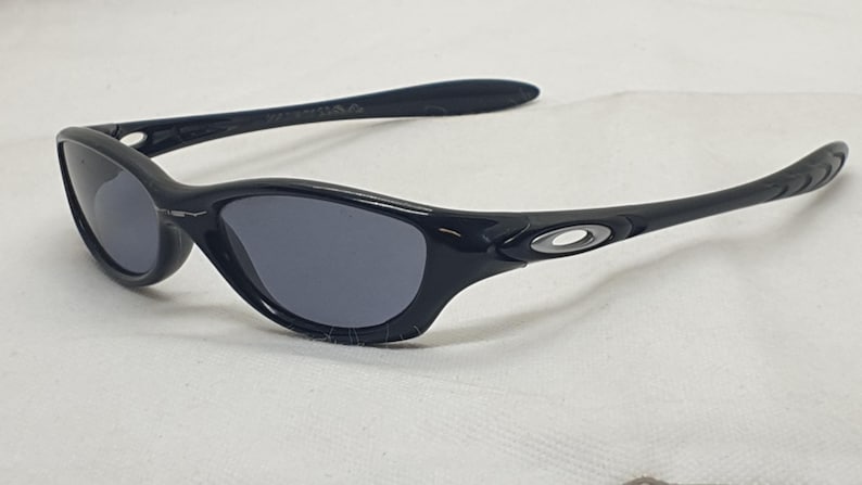 Puede incluir: Gafas de sol negras con lentes grises. Las gafas tienen una montura y patillas curvas, con un peque&ntilde;o emblema plateado en las patillas. Las lentes son de color gris oscuro y la montura es negra brillante.
