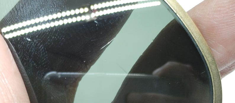 May include: Close-up of sunglasses with a gold-colored frame and green-tinted lenses. The lenses have a slight scratch. The sunglasses are held by a person's fingers.